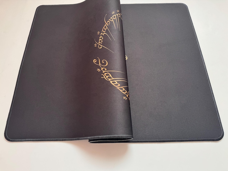 Puede incluir: Una alfombrilla de rat&oacute;n rectangular negra con un dise&ntilde;o dorado de escritura &eacute;lfica. La alfombrilla est&aacute; parcialmente enrollada, revelando la parte inferior. Los bordes est&aacute;n cosidos con hilo negro. El dise&ntilde;o es de El Se&ntilde;or de los Anillos.