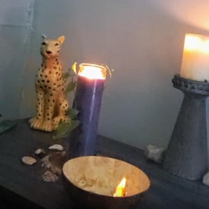 Peut inclure: Une composition décorative avec une figurine de guépard en céramique, une bougie violette allumée, une bougie allumée dans une coquille de noix de coco et une grande bougie blanche sur un piédestal gris. La scène est éclairée par la lueur chaude des flammes.