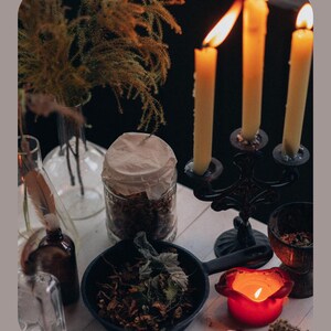 May include: A still-life arrangement featuring lit candles in a black candelabra, a small cast iron pan with herbs, and a red candle holder. Other elements include glass bottles, a jar with a paper cover, and dried plants, creating a mystical aesthetic.