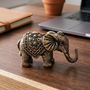 Pode incluir: Uma detalhada estatueta de elefante dourado em estilo antigo com padrões intrincados nas costas e orelhas. O elefante tem uma tromba enrolada e está posicionado sobre uma superfície de madeira, sugerindo um item decorativo ou de colecionador.