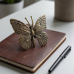 May include: A brass-toned metal butterfly figurine with detailed wing patterns and two antennae. The butterfly has a textured surface with dark recessed areas and lighter raised edges, creating a metallic finish. It is positioned upright.