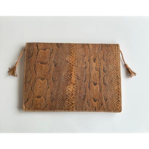 May include: A rectangular clutch bag featuring a textured brown and tan snakeskin pattern. The bag has a flat, envelope-style design with thin, braided side cords that terminate in small, matching tassels on both the left and right sides.