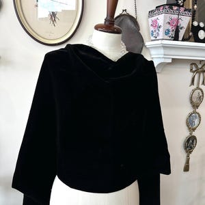 May include: A black velvet cape with a cowl neck and long sleeves, displayed on a mannequin. The cape is the focal point, with a framed botanical print and other decorative items in the background. The cape is made of a soft, luxurious fabric.