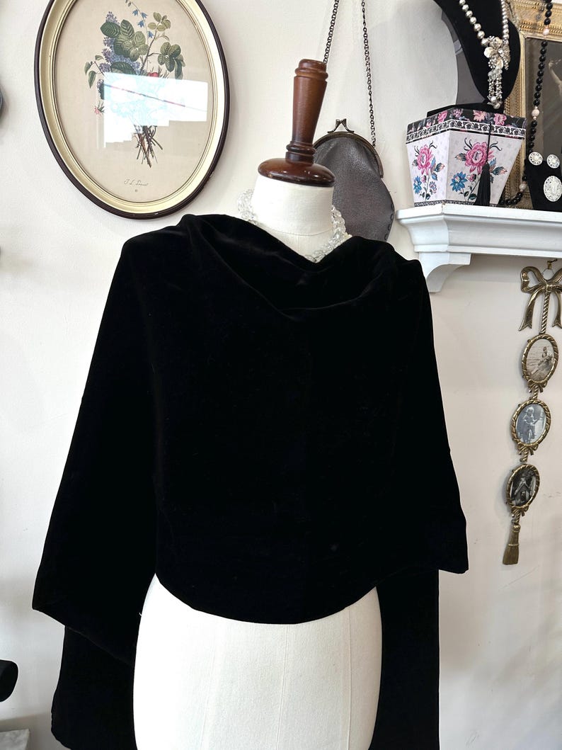 May include: A black velvet cape with a cowl neck and long sleeves, displayed on a mannequin. The cape is the focal point, with a framed botanical print and other decorative items in the background. The cape is made of a soft, luxurious fabric.