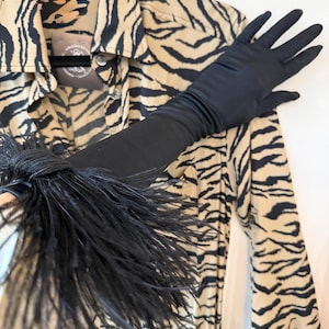 May include: A long black satin glove featuring a dense black feather trim at the wrist, positioned over a tan button-down shirt with a black tiger stripe pattern. The shirt includes a visible circular brand tag near the collar area.