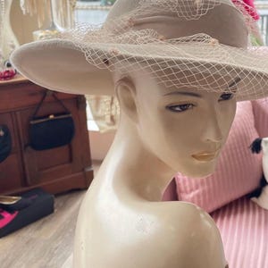 May include: A beige felt hat with a wide brim and a delicate veil. The hat is adorned with small, pearl-like embellishments.
