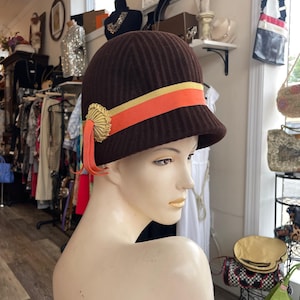Puede incluir: Un sombrero cloche marrón con textura acanalada, adornado con una cinta dorada y naranja. El sombrero presenta un adorno dorado y se exhibe en un maniquí en una tienda vintage.