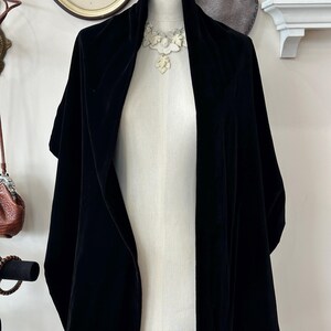 May include: A black velvet shawl displayed on a mannequin. The shawl features a broad collar and hangs down the front. A white floral necklace is seen at the neckline. The mannequin is on a metal stand.