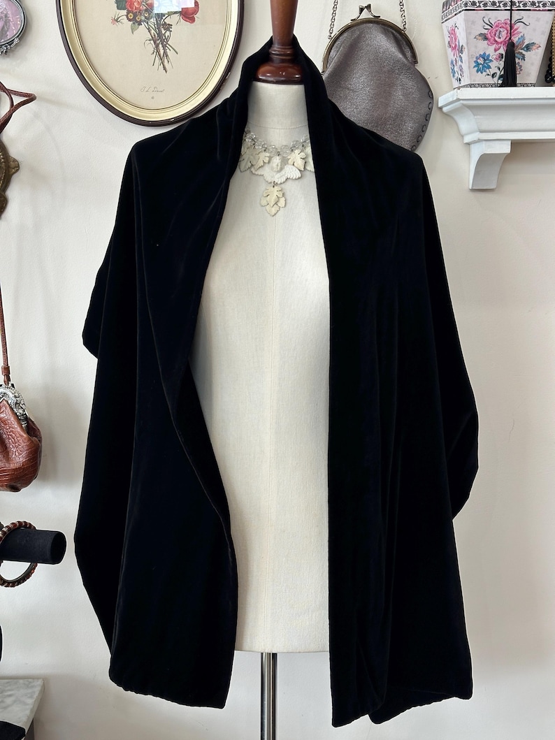 May include: A black velvet shawl displayed on a mannequin. The shawl features a broad collar and hangs down the front. A white floral necklace is seen at the neckline. The mannequin is on a metal stand.