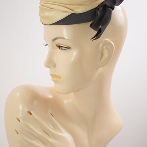 May include: A beige and brown satin hat with a black ribbon bow. The hat is worn on a mannequin's head.