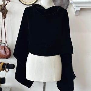 May include: A black velvet cape with a cowl neck is displayed on a mannequin. The cape has wide sleeves and a flowing design. The fabric appears soft and drapes elegantly. The background includes vintage decor.
