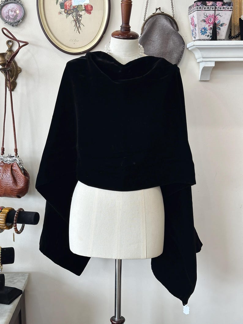 May include: A black velvet cape with a cowl neck is displayed on a mannequin. The cape has wide sleeves and a flowing design. The fabric appears soft and drapes elegantly. The background includes vintage decor.