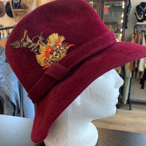 May include: A burgundy felt fedora hat with a wide brim and a decorative feather accent. The hat has a black ribbon around the crown.