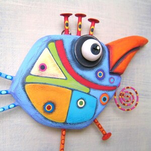 May include: A whimsical, hand-painted bird sculpture in shades of blue, orange, red, yellow, and green. The bird has a large eye, a prominent orange beak, and decorative accents. The sculpture is mounted on a light gray surface.