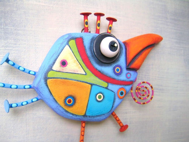 May include: A whimsical, hand-painted bird sculpture in shades of blue, orange, red, yellow, and green. The bird has a large eye, a prominent orange beak, and decorative accents. The sculpture is mounted on a light gray surface.
