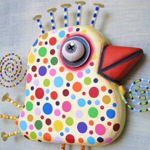 May include: A whimsical, hand-painted bird sculpture with a cream-colored body adorned with colorful polka dots. It features a red beak, a large eye, and decorative spirals. The bird has nail-like accents on its head, creating a unique, folk art style.