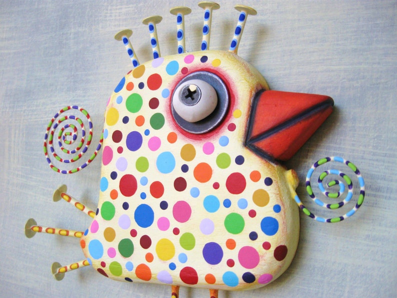 May include: A whimsical, hand-painted bird sculpture with a cream-colored body adorned with colorful polka dots. It features a red beak, a large eye, and decorative spirals. The bird has nail-like accents on its head, creating a unique, folk art style.