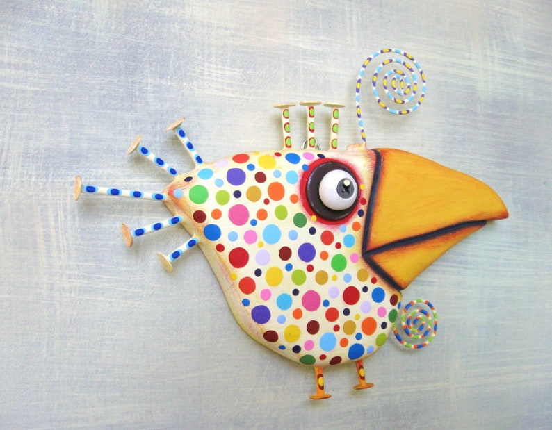 May include: Whimsical bird sculpture with a large yellow beak and a body covered in colorful polka dots. The bird has a large, expressive eye and decorative wire accents. The sculpture is mounted on a light blue background.