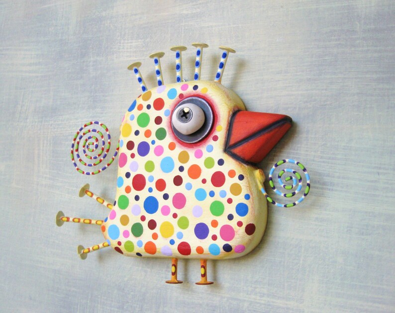 May include: A whimsical, hand-painted bird sculpture with a cream-colored body covered in colorful polka dots. It features a red beak, a large eye, and decorative spiral accents. The bird has six yellow legs and blue-tipped head spikes.