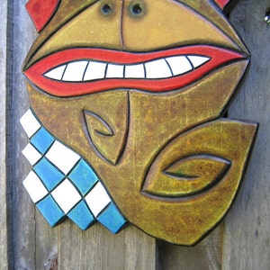 May include: A carved wooden mask with a smiling face. The mask features a red mouth with white teeth, a brown face, and a blue and white diamond-patterned collar. The mask is likely for decorative purposes.
