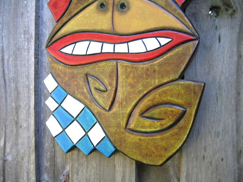 May include: A carved wooden mask with a smiling face. The mask features a red mouth with white teeth, a brown face, and a blue and white diamond-patterned collar. The mask is likely for decorative purposes.
