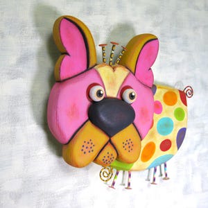 Puede incluir: Una colorida y caprichosa decoraci&oacute;n de pared con forma de perro. La cara del perro es principalmente rosa con detalles amarillos y negros, ojos grandes y una nariz gris oscuro. El cuerpo es amarillo con lunares grandes y coloridos. Montado en una pared blanca.