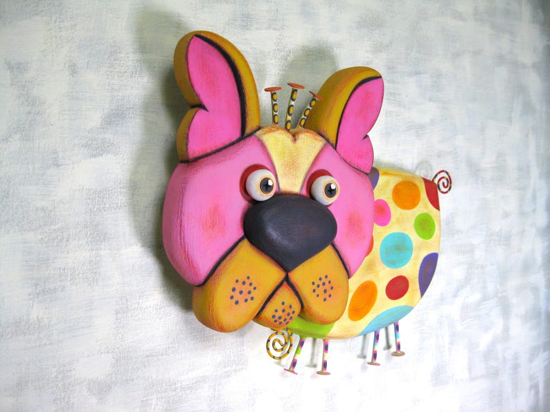 Puede incluir: Una colorida y caprichosa decoraci&oacute;n de pared con forma de perro. La cara del perro es principalmente rosa con detalles amarillos y negros, ojos grandes y una nariz gris oscuro. El cuerpo es amarillo con lunares grandes y coloridos. Montado en una pared blanca.