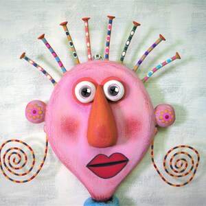 May include: A pink face with a large orange nose, white eyes, and red lips. The face has a colourful, spiky hairdo and two spiral-shaped earrings.
