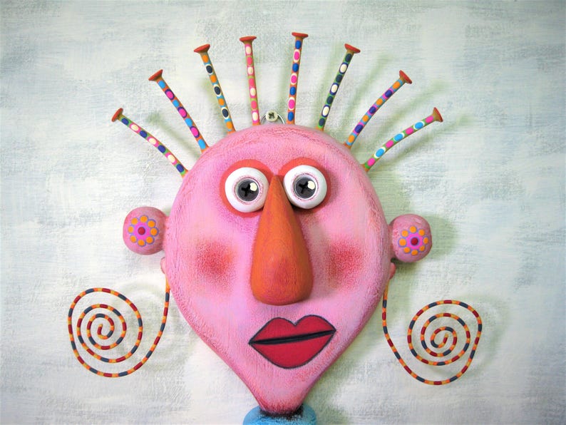 May include: A pink face with a large orange nose, white eyes, and red lips. The face has a colourful, spiky hairdo and two spiral-shaped earrings.