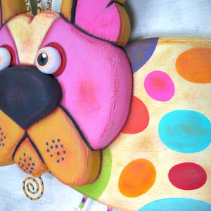 Puede incluir: Escultura de perro de madera caprichosa y colorida con una gran nariz negra, orejas rosas y detalles en naranja y amarillo. El cuerpo del perro est&aacute; decorado con grandes lunares vibrantes en varios colores. La escultura tiene un dise&ntilde;o caricaturesco y juguet&oacute;n.