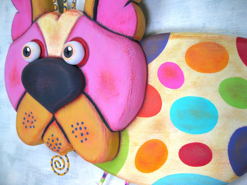 Puede incluir: Escultura de perro de madera caprichosa y colorida con una gran nariz negra, orejas rosas y detalles en naranja y amarillo. El cuerpo del perro est&aacute; decorado con grandes lunares vibrantes en varios colores. La escultura tiene un dise&ntilde;o caricaturesco y juguet&oacute;n.