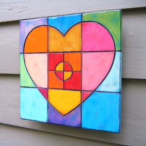 May include: A colorful heart-shaped mosaic made of ceramic tiles. The heart is composed of various shades of pink, orange, yellow, blue, green, and red. The center of the heart features a red circle with a yellow center and a black cross.