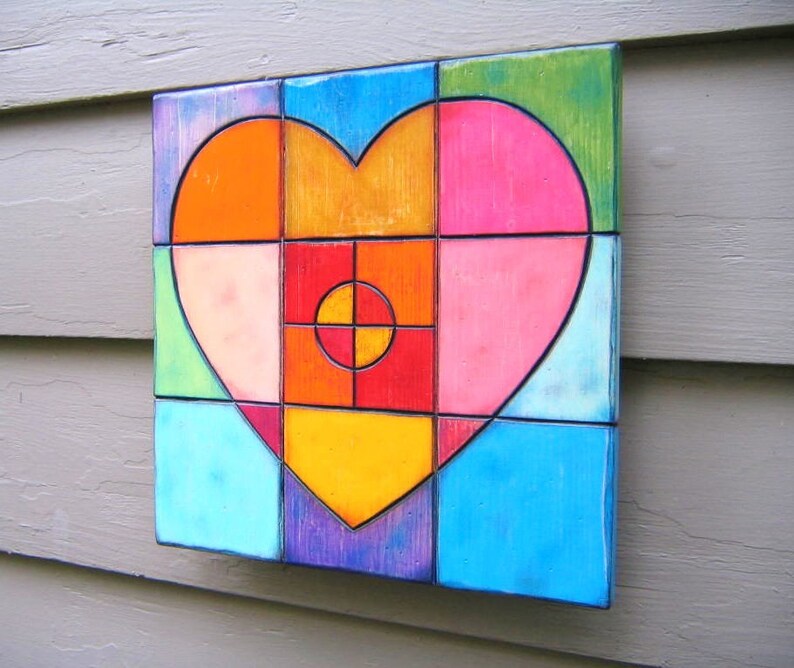 May include: A colorful heart-shaped mosaic made of ceramic tiles. The heart is composed of various shades of pink, orange, yellow, blue, green, and red. The center of the heart features a red circle with a yellow center and a black cross.
