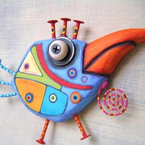 May include: A whimsical, colorful bird sculpture with a large orange beak and a blue body. The bird features a large eye, red and yellow accents, and a spiral tail. The design incorporates geometric shapes and a variety of colors, including blue, orange, yellow, and green.