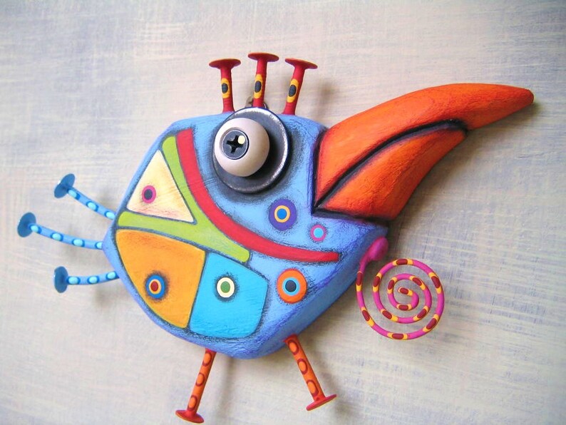 May include: A whimsical, colorful bird sculpture with a large orange beak and a blue body. The bird features a large eye, red and yellow accents, and a spiral tail. The design incorporates geometric shapes and a variety of colors, including blue, orange, yellow, and green.