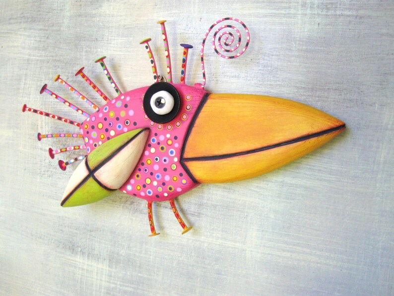 May include: A whimsical pink and orange bird sculpture with a long beak, black eye, and a spiral antenna. The bird is decorated with colorful polka dots and has a green wing.