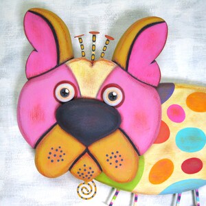 Puede incluir: Escultura de perro de madera caprichosa con cara rosa, detalles amarillos y naranjas y nariz negra. El cuerpo del perro est&aacute; decorado con lunares de colores. La escultura tiene un estilo de arte popular juguet&oacute;n.