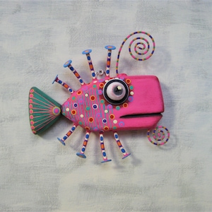 May include: A whimsical pink fish sculpture with a large eye, colorful polka dots, and a spiral tail. The fish has a green and pink tail fin and multiple blue and orange spikes.