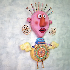 May include: A whimsical, handmade sculpture of a pink-faced figure with large eyes, a long orange nose, and a red mouth. The figure has yellow wings, a colourful patterned body, and a blue and white striped collar. The sculpture is made of various materials and is painted in bright colours.