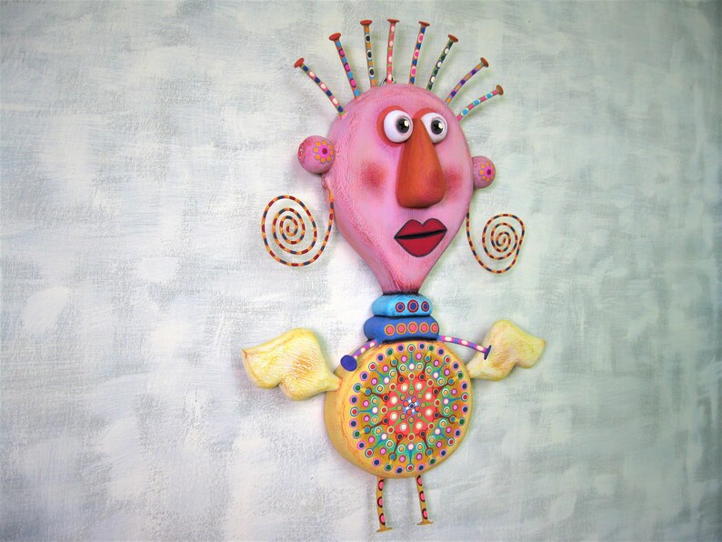 May include: A whimsical, handmade sculpture of a pink-faced figure with large eyes, a long orange nose, and a red mouth. The figure has yellow wings, a colourful patterned body, and a blue and white striped collar. The sculpture is made of various materials and is painted in bright colours.
