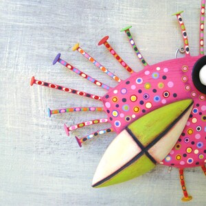 May include: A whimsical pink bird sculpture with a green beak and black eye. The bird has a colorful polka dot pattern and is decorated with colorful wooden sticks.