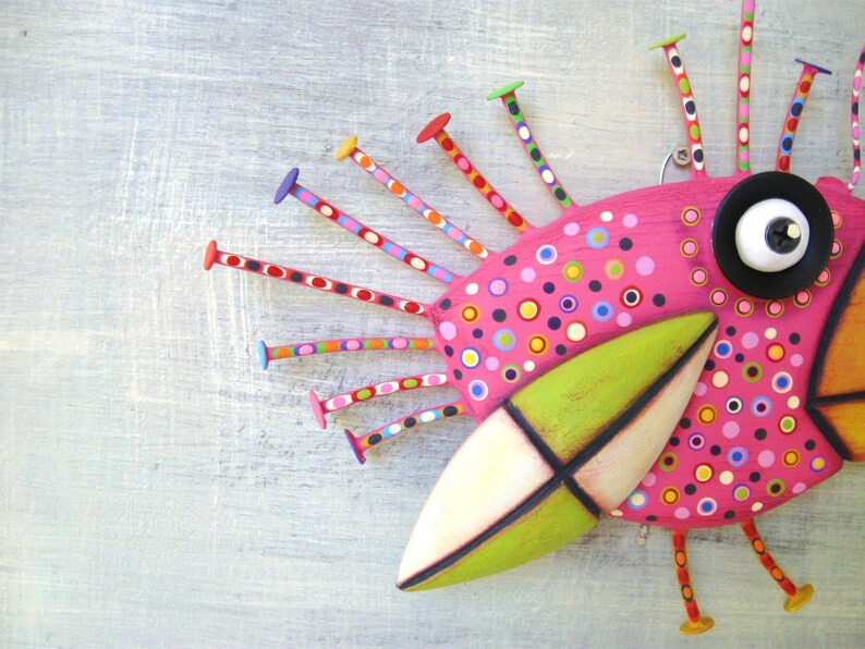 May include: A whimsical pink bird sculpture with a green beak and black eye. The bird has a colorful polka dot pattern and is decorated with colorful wooden sticks.
