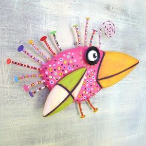 May include: A whimsical pink bird sculpture with a long orange beak, green wings, and a colorful, polka-dotted body. The bird has a large black eye with a white pupil and a spiral antenna on its head.