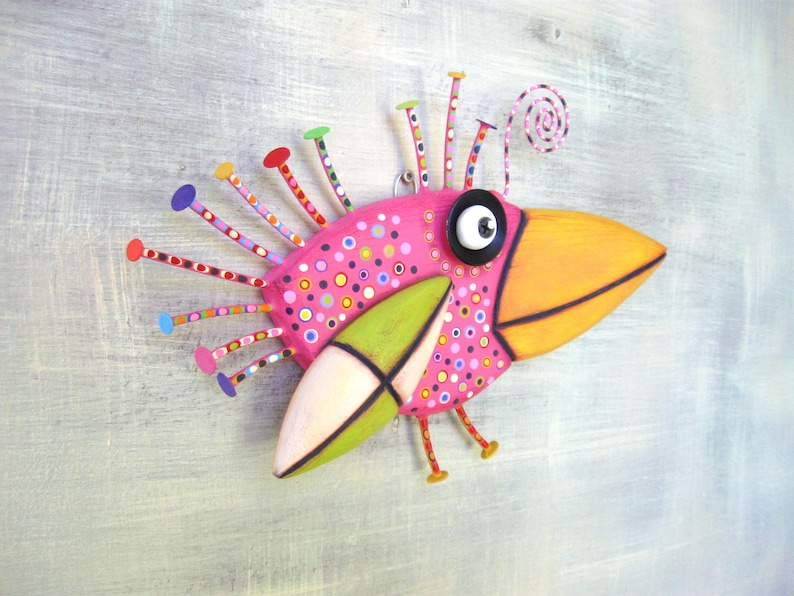May include: A whimsical pink bird sculpture with a long orange beak, green wings, and a colorful, polka-dotted body. The bird has a large black eye with a white pupil and a spiral antenna on its head.