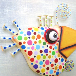 May include: Whimsical, colorful bird sculpture with a large yellow beak and a body covered in multi-colored polka dots. The bird has a large eye, and decorative elements including a spiral and stick-like appendages.