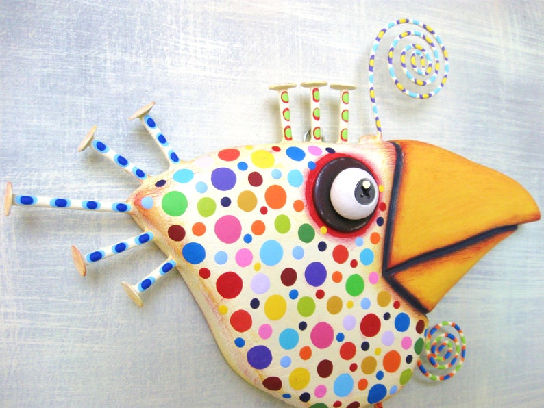 May include: Whimsical, colorful bird sculpture with a large yellow beak and a body covered in multi-colored polka dots. The bird has a large eye, and decorative elements including a spiral and stick-like appendages.