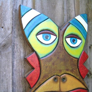 May include: A colorful, hand-painted wooden mask with a stylized face. The mask features bright blue eyes, a yellow and green face, and red accents. The top of the mask has blue and white stripes. The mask is mounted on a weathered wooden background.