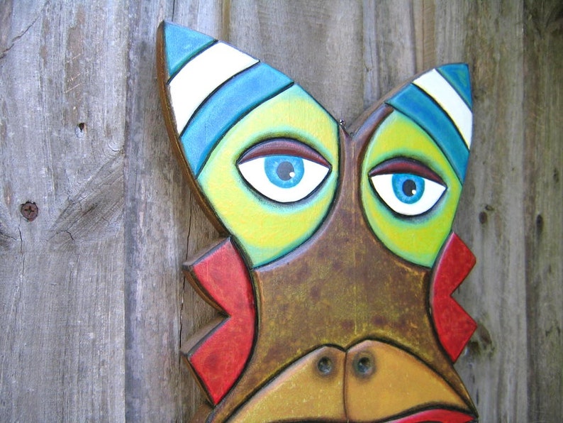 May include: A colorful, hand-painted wooden mask with a stylized face. The mask features bright blue eyes, a yellow and green face, and red accents. The top of the mask has blue and white stripes. The mask is mounted on a weathered wooden background.