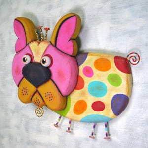 Puede incluir: Escultura de perro de madera pintada a mano. El perro presenta una cara rosa, orejas amarillas y un cuerpo adornado con lunares coloridos. Tiene una nariz negra, ojos grandes y una cola rizada. La escultura est&aacute; montada sobre una superficie gris claro.