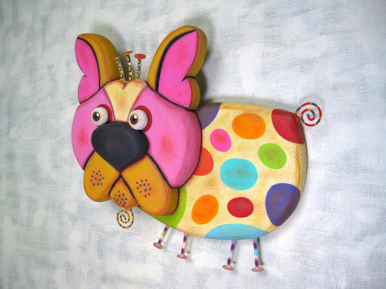 Puede incluir: Escultura de perro de madera pintada a mano. El perro presenta una cara rosa, orejas amarillas y un cuerpo adornado con lunares coloridos. Tiene una nariz negra, ojos grandes y una cola rizada. La escultura est&aacute; montada sobre una superficie gris claro.